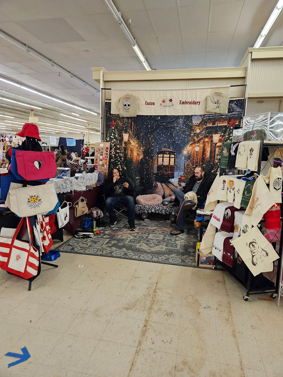 Two people sit in a cozy embroidery booth, surrounded by bags and textiles. A backdrop shows a snowy street scene. Sign reads "Custom Embroidery."