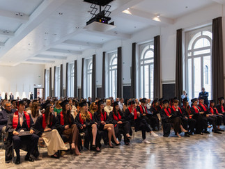 Graduates in black gowns and red sashes seated in rows during the ISB Class of 2026 graduation ceremony at Palazzo de' Toschi, Bologna. The elegant hall features tall arched windows and a checkered marble floor.