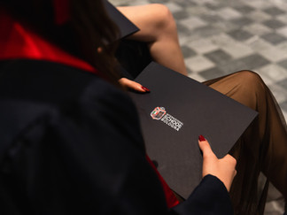 Close-up of a graduate's hands holding a black diploma folder embossed with the International School of Bologna logo, wearing a black gown with a red sash.