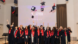 Graduates of the International School of Bologna Class of 2026 celebrate by tossing their black mortarboard caps with red tassels into the air. The group of approximately twenty students, dressed in black graduation gowns and red sashes, stand in a row with arms raised and joyful expressions. Behind them, a projected screen displays the school's logo and the text 'International School of Bologna – Class of 2026.