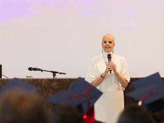 A woman in a white outfit speaks into a microphone on stage during the ISB graduation ceremony, with graduates in caps and gowns visible in the foreground.