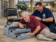 Mall staff comforts an older man in a maroon polo as he lies beside the ResQUp fall recovery device, looking tired and distressed.