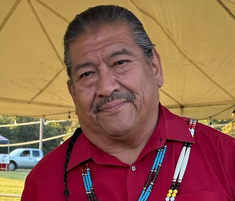 David Bible wearing a red shirt with beaded necklaces, standing under a tan tent.