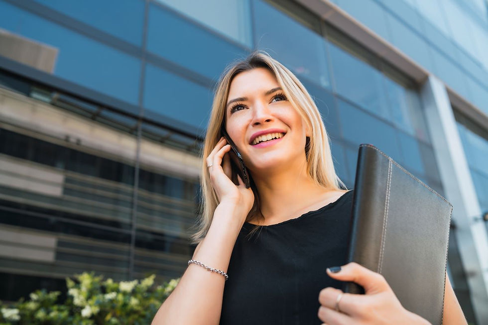 mulher sorridente com cabelos loiros está ao telefone, segurando um caderno preto, com um prédio de escritórios ao fundo.