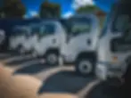a row of Isuzu trucks parked outside Truckrite's facility under a clear blue sky with trees in the background.