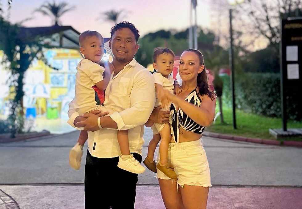 Smiling parents each hold a young child outdoors at dusk, with blurred trees and colorful lights in the background.