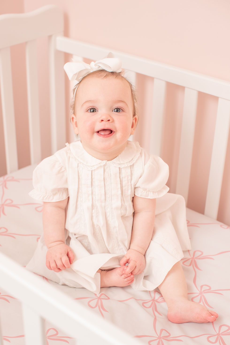 Baby girl sitting in white crib in pink nursery wearing a white dress and smiling during her nine month milestone portrait session in her home near Benson, NC.