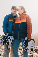 A couple holds their helmets with the sand dunes behind them