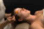Person lying down receiving a facial treatment with a brush. Calm atmosphere, dark background, white cream on forehead, orange brush.