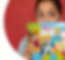 Shay Abigail peeks over the book cover of her debut book, My Name Is Maya. Her dark black hair is pulled back in a bun. Eyebrows up and looking right with her brown eyes. The book cover shows a blue sky, and the character Maya stands front and center, wearing a blue cape, big red glasses, and a bold smile. She has her foot leaning on a blue elephant, one hand on her hip and the other raised high in a fist. Her dad is in orange shorts and a green/blue shirt to her right. He is blond and wearing big brown glasses. To the right of Dad sits a white dog, a happy husky pet wearing a red collar. To the left of Maya is her mom, with red hair pulled back in a bun, a purple tank top, and orange shorts. She sits on a sky-blue blanket. In front of the mother sits a bald baby/toddler wearing a light blue shirt. All eyes are on Maya.