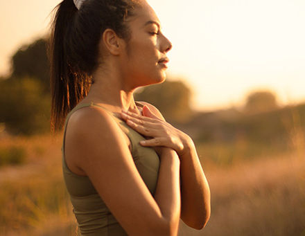 Woman standing in a field. Her eyes are closed and her hands are placed on her heart