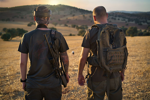 Two Hunters With Backpacks Standing In Field At Sunset