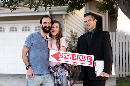 Couple With Real Estate Agent Holding Open House Sign In Front Of Home