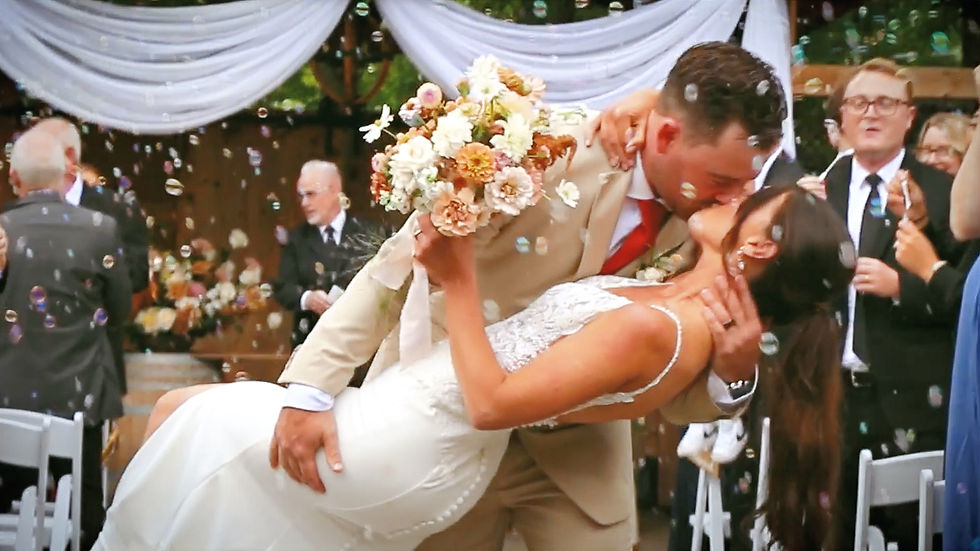 Bride and groom kiss in a bubble-filled garden, bride holding bouquet, groom in beige suit. Joyful wedding scene with guests.