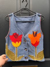 A blue denim waistcoat appliqued with a red and orange tulip. There is an orange blank stitch around the cuffs.