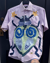 a light purple gingham shirt with an appliqué beetle embroidered onto it. the beetle has a colour scheme of green, blue, and yellow. there are two blue spirals and a blue heart on the outer shell of the beetle. the beetle also has star shaped antenna and light blue eyes.
