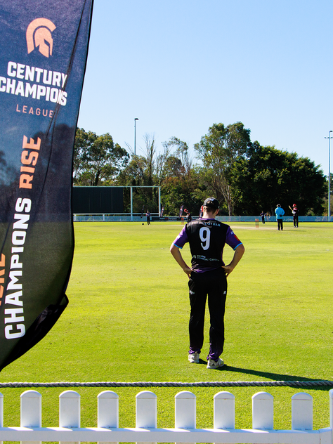 Cricket player in black and purple uniform stands on the field during a Century Champions League match, with a flag reading 'Where Champions Rise' in the foreground and other players positioned in the background.