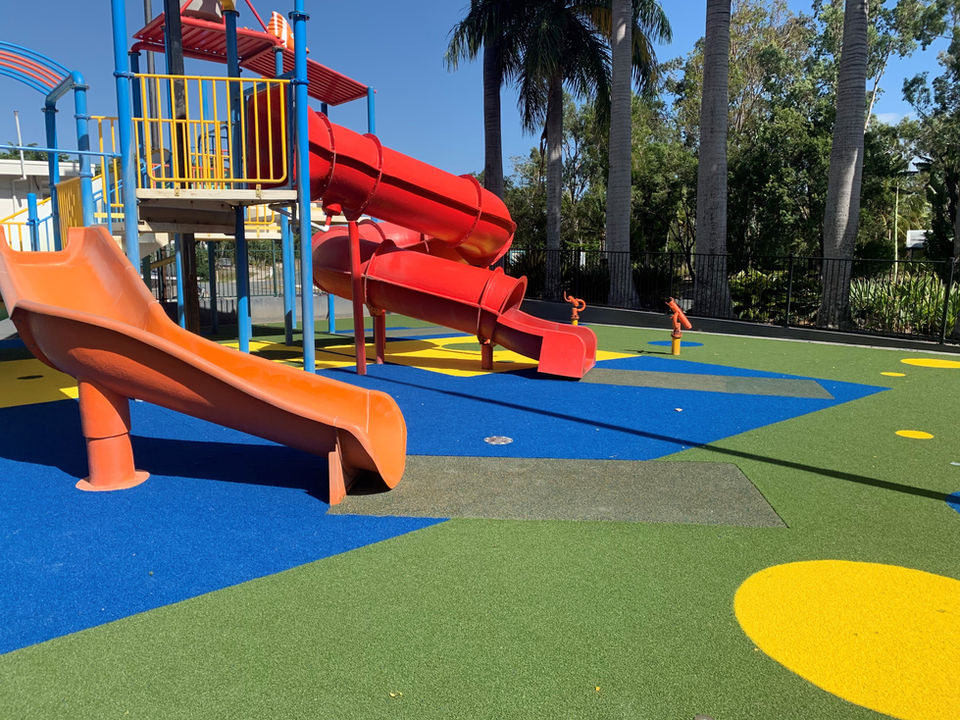 A colourful outdoor playground, featuring vibrant green synthetic turf with various blue and yellow shapes. The area is surrounded by palm trees.
