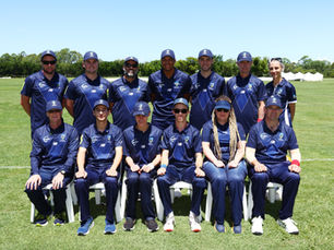 Group of cricket officials in navy uniforms posing for a team photo on a grass field.