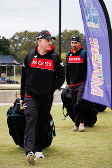 Two cricket players in black and red uniforms walking with large black bags..