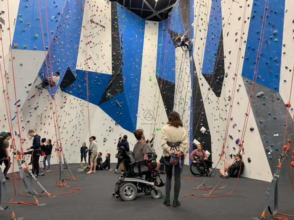 [Image ID: an indoor rock climbing venue with various people, some are making their way up the wall and others are supporting them from the ground. In the middle of the area there is a young person using a wheelchair and another person standing next to them.]