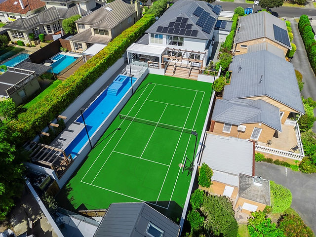 Aerial view of a residential tennis court made with bright green synthetic turf, in a modern backyard. Surrounding the tennis court is a lap pool, concrete fencing and greenery.