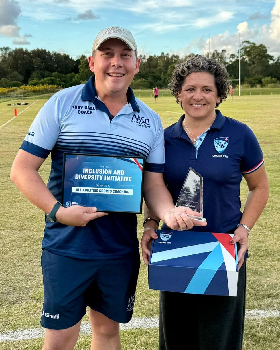 Smiling pair on a grassy sports field holding award plaque and trophy; plaque reads Inclusion and Diversity Initiative.