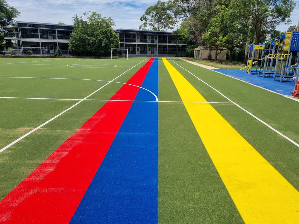 A large outdoor oval with sports field markings. Along the side of the oval are vibrant synthetic grass stripes in red, blue, green and yellow running in a straight line.