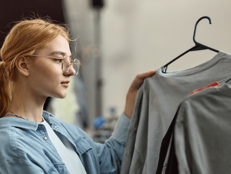 Young woman with glasses browses gray shirts on hangers in a clothing store, calmly examining the rack.