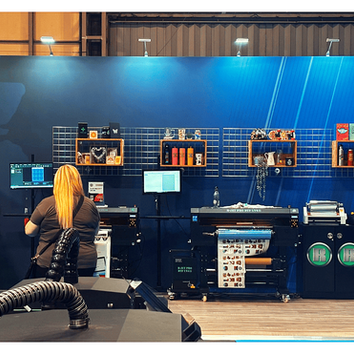 Two people view large printers and product displays in a blue print shop booth with R logos and monitors.