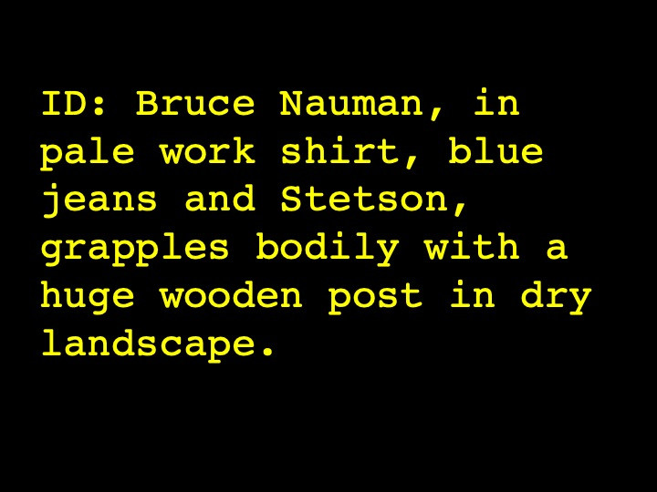 Bruce Nauman, in pale work shirt, blue jeans and Stetson, grapples bodily with a huge wooden post in dry landscape.