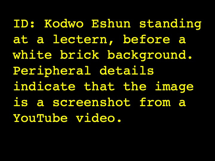 Kodwo Eshun standing at a lectern, before a white brick background. Peripheral details indicate that the image is a screenshot from a YouTube video