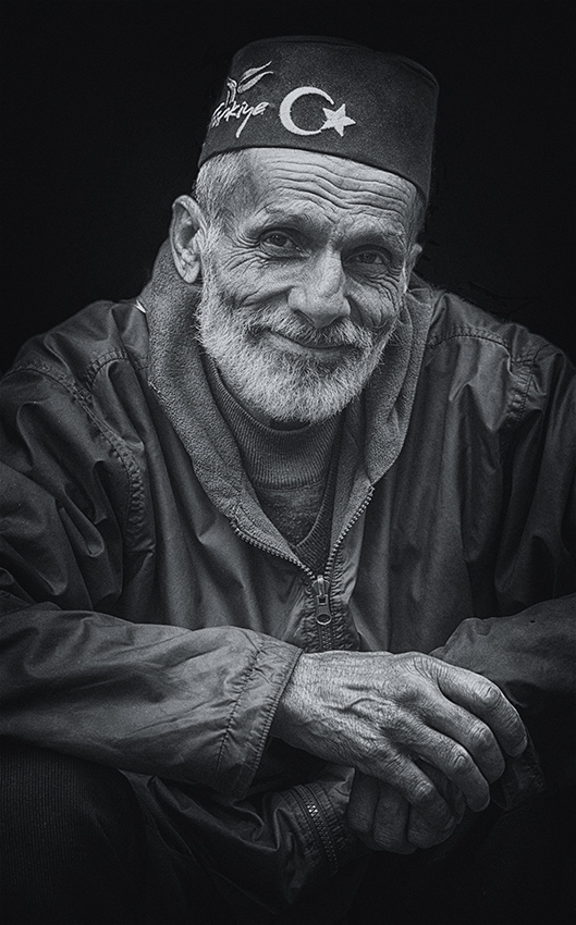 Black and white portrait of an elderly man wearing a traditional fez hat with a crescent and star emblem, smiling gently — a dignified cultural portrait by Cristian Mitrani.