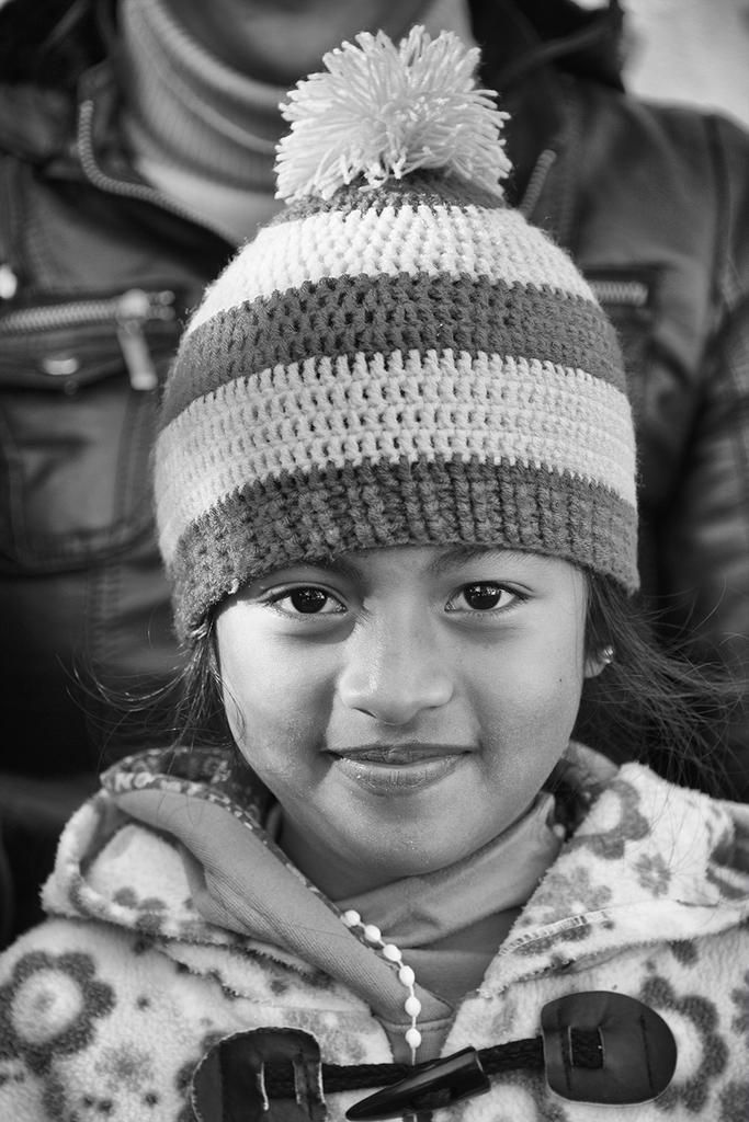 Black and white portrait of a smiling young girl wearing a striped woolen hat and floral jacket — a heartwarming scene by Cristian Mitrani.
