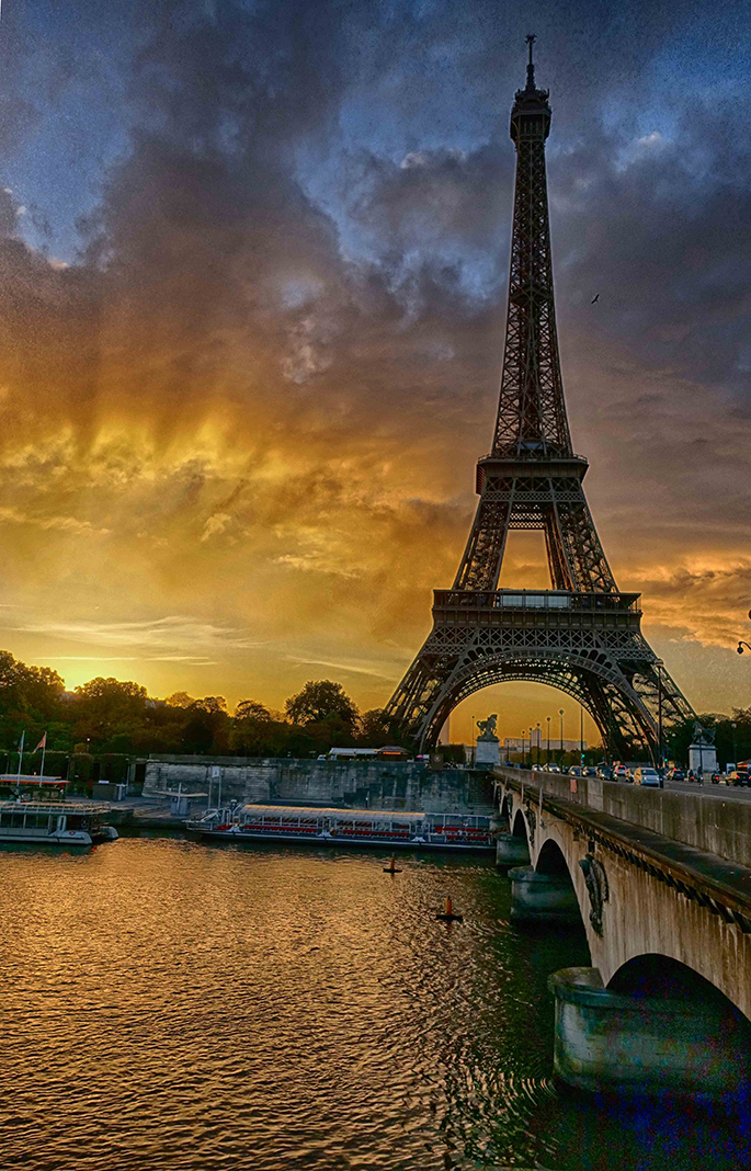 Photograph by Cristian Mitrani depicting the Eiffel Tower in Paris at sunset, with a dramatic sky filled with dark clouds illuminated by the golden and orange hues of the setting sun. The scene includes the River Seine in the foreground with boats and a bridge, capturing the iconic Parisian skyline under a striking sky.