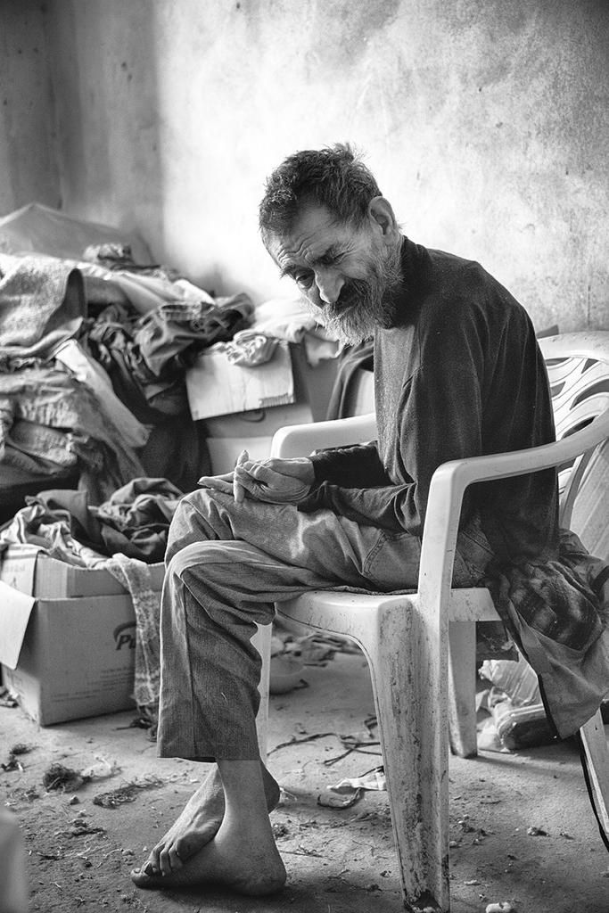Black and white photo of an elderly man sitting barefoot on a plastic chair in a cluttered room, captured in a moment of quiet reflection — by Cristian Mitrani.