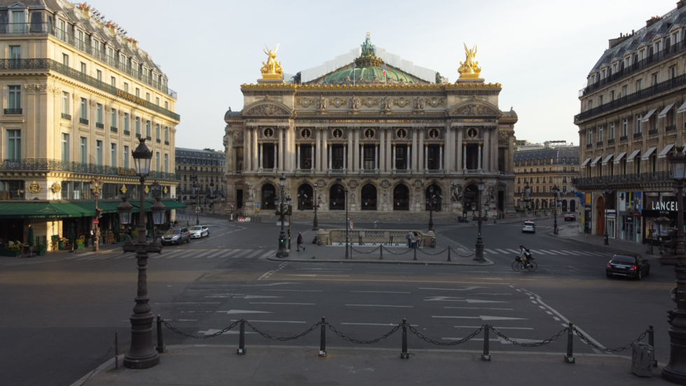 An image taken by Cristian Mitrani depicting the Palais Garnier (Opera House) in Paris. The grand historic building features intricate architectural details, with a richly decorated facade and sculptures. The photo captures the wide front plaza with a few parked vehicles and pedestrians, under a clear sky, showcasing Mitrani's signature style of detailed and textured photography.