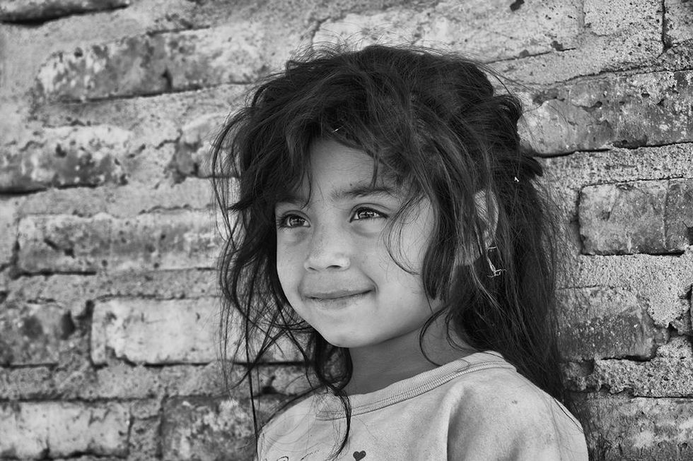 Black and white portrait of a smiling young girl with tousled hair standing against a brick wall — a tender human moment documented by Cristian Mitrani.