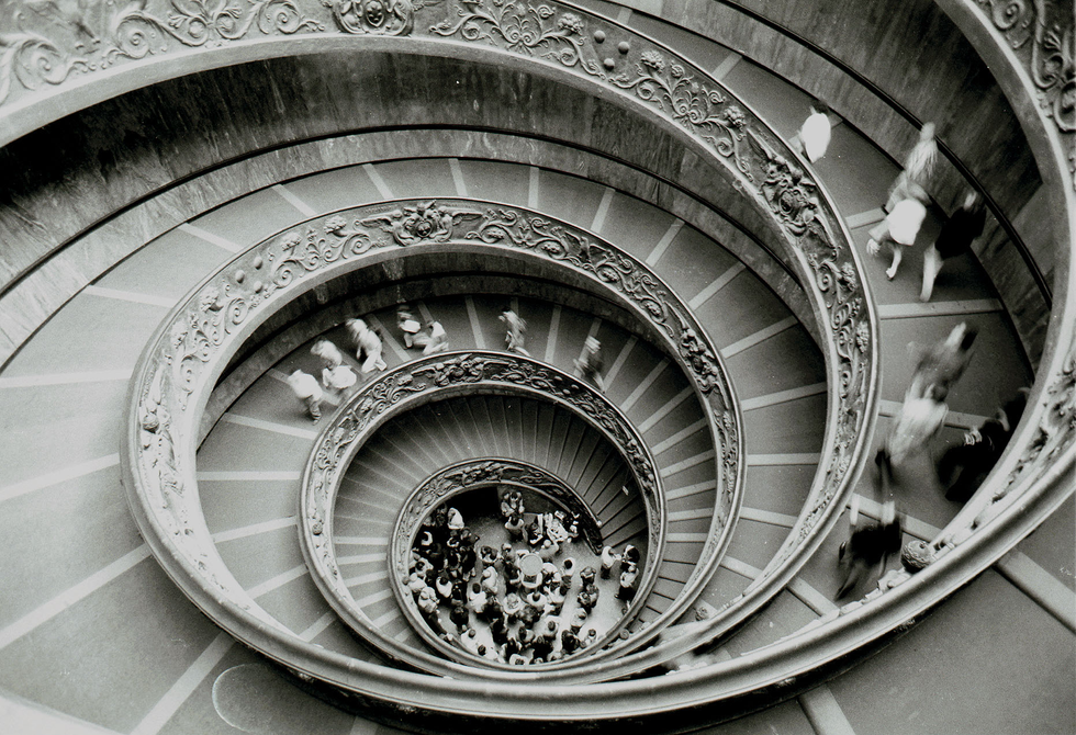 Black-and-white aerial photograph by Cristian Mitrani showing a spiraling staircase viewed from above. The staircase curves downward in a tight spiral, revealing intricate architectural details along the railings and steps. At the bottom, a group of people and small objects are visible, emphasizing the depth and scale of the spiral design. The image captures Mitrani's signature style of atmospheric urban and architectural photography, highlighting the elegance and complexity of the spiral structure.