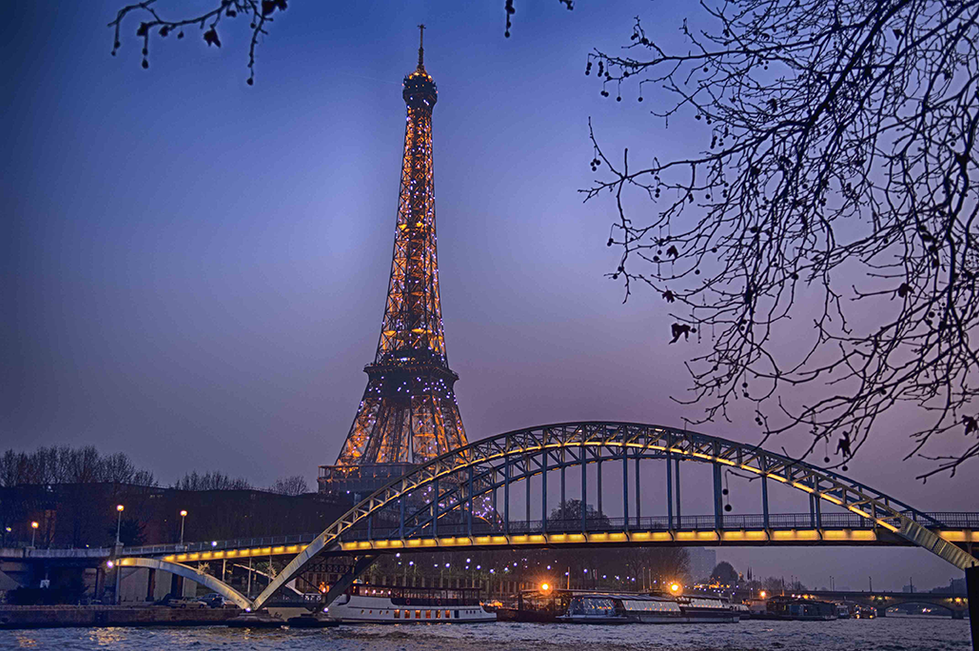 Photograph by Cristian Mitrani showcasing the illuminated Eiffel Tower in Paris at dusk, with its intricate iron lattice structure glowing against the twilight sky. In the foreground, a gracefully arched bridge spans the river, with reflections shimmering on the water below. Bare tree branches frame the scene, adding depth and a sense of seasonality to the stunning cityscape.