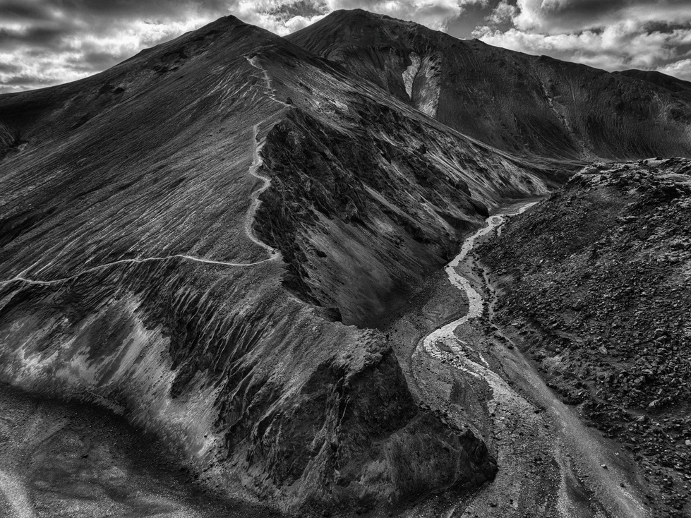 A black-and-white photograph by Cristian Mitrani capturing a dramatic mountain landscape with steep, rugged slopes. A winding trail cuts through the terrain, leading the viewer’s eye along the contours of the mountain and emphasizing its imposing scale and textured surface.