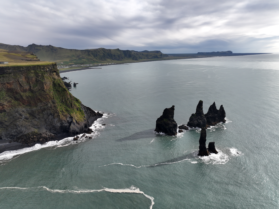 A panoramic view of a rugged coastline with steep cliffs and dark volcanic rock formations rising from the ocean, under a cloudy sky. The scene's dramatic natural beauty and striking contrasts reflect Cristian Mitrani's artistic focus on capturing powerful landscapes and raw nature.