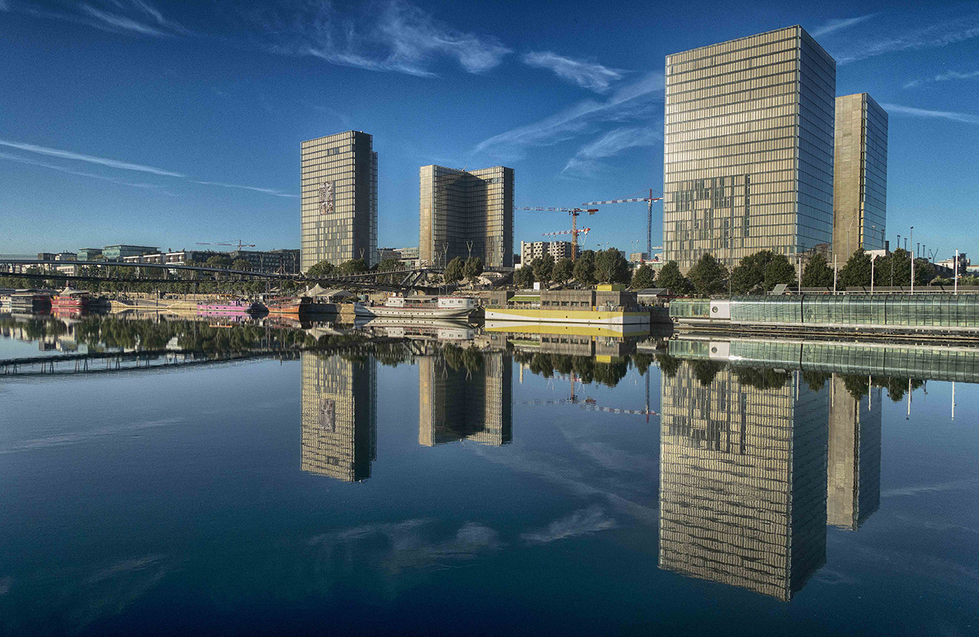 A photograph by Cristian Mitrani depicting a modern cityscape with three tall, rectangular office buildings reflected in a calm river. The sky is clear with a few wispy clouds, and construction cranes are visible in the background, indicating ongoing development. The water creates a mirror-like reflection of the buildings, emphasizing the urban environment and architectural design.