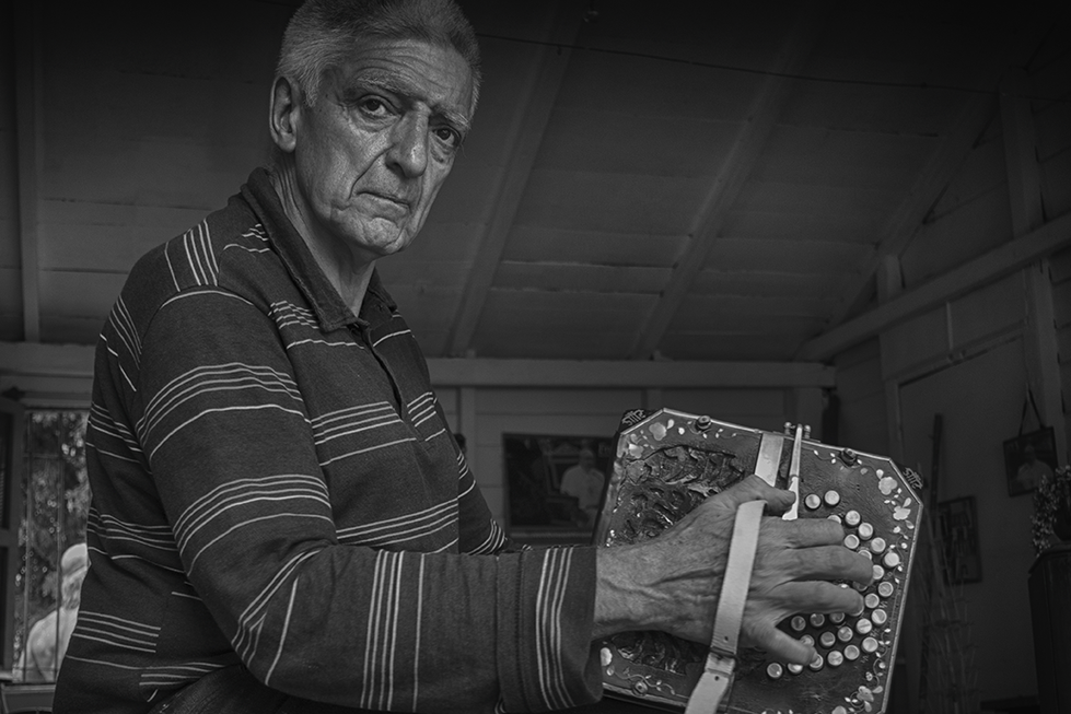 Black and white portrait of an elderly man playing a traditional accordion, seated indoors with a focused expression — a cultural moment captured by Cristian Mitrani.
