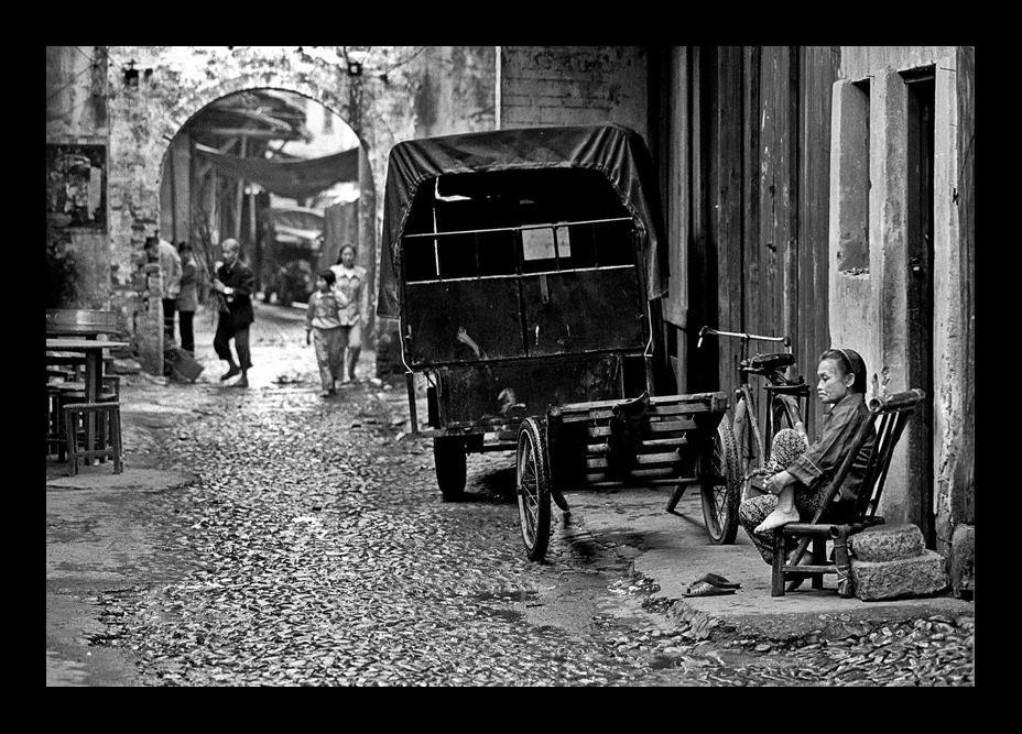 Black-and-white photograph by Cristian Mitrani depicting a cobblestone street scene with a horse-drawn carriage parked beside a building. A woman sits on a bench nearby, while a group of people walk in the background. The scene captures everyday life in a historic town or city, with the carriage and street setting adding a nostalgic atmosphere.