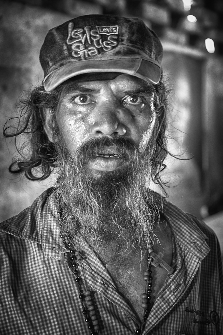 Black and white portrait of a man with a long beard and expressive eyes, wearing a cap with script, conveying intensity and life stories — a powerful study by Cristian Mitrani.