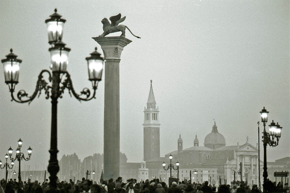 Black-and-white photograph by Cristian Mitrani capturing a historic city square featuring a tall column topped with a lion sculpture. In the background, the bell tower of St. Mark's Basilica and other classical architecture are visible, shrouded in a misty or foggy atmosphere. The scene reflects Mitrani's atmospheric urban photography style, emphasizing timeless architectural elements and moody ambiance.