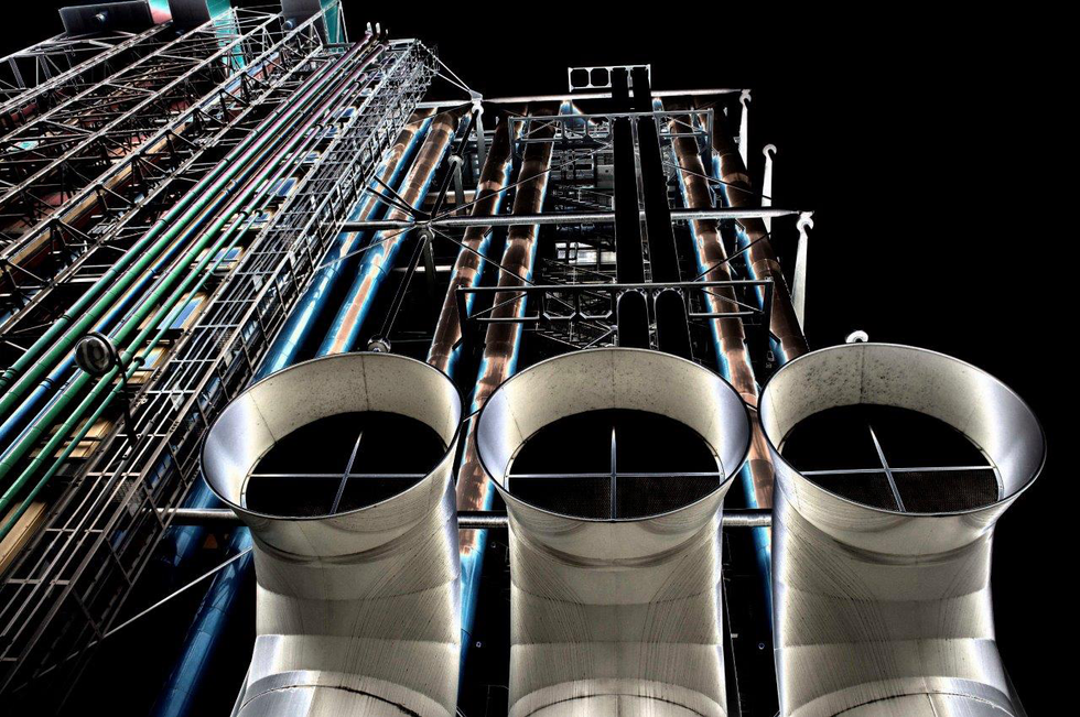 Photograph by Cristian Mitrani showing an upward view of pompidou against a dark night sky. The image features large, curved white pipes in the foreground, with an intricate network of metal pipes and structures illuminated by artificial lighting, highlighting the complexity and scale of the industrial infrastructure.