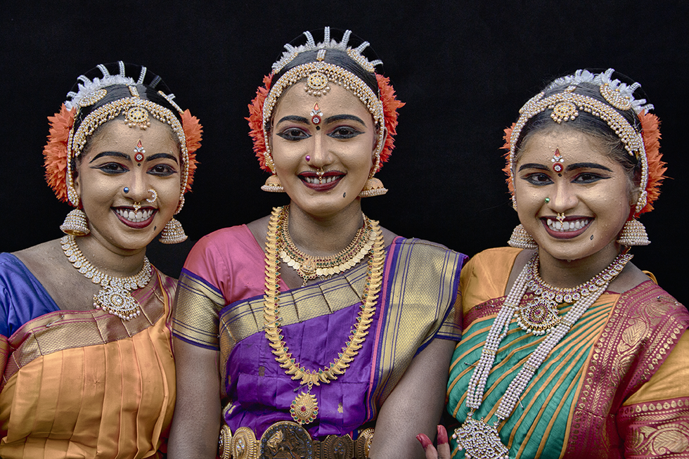 Three smiling women dressed in traditional Indian attire with elaborate jewelry and headpieces, posing together against a black background — a vibrant cultural portrait by Cristian Mitrani.