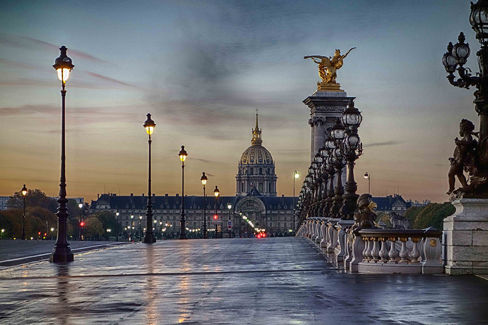 Photograph by Cristian Mitrani of the Alexandre III Bridge in Paris during early evening, featuring the ornate golden statues atop the bridge's pillars. The scene shows the illuminated lamps along the bridge, with the domed Les Invalides building in the background against a moody, cloudy sky, and the wet surface of the bridge reflecting the lights.